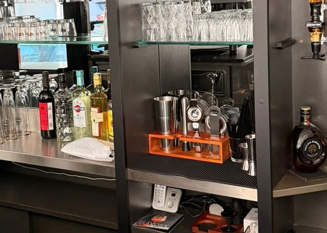 Die Arbeitsseite der Bar: Flaschen mit Martini Bianco, Grappa und Rotwein auf der Edelstahlablage, daneben Rührglas, Shaker und Barsieb im orangefarbenen Halter, darüber Gläser im Regal.