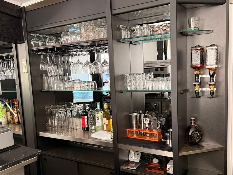 Das Rückbuffet hinter der Theke: Gläser in Reihen auf Glasböden, hängende Weingläser, Flaschen und das Barwerkzeug auf der Edelstahlablage.