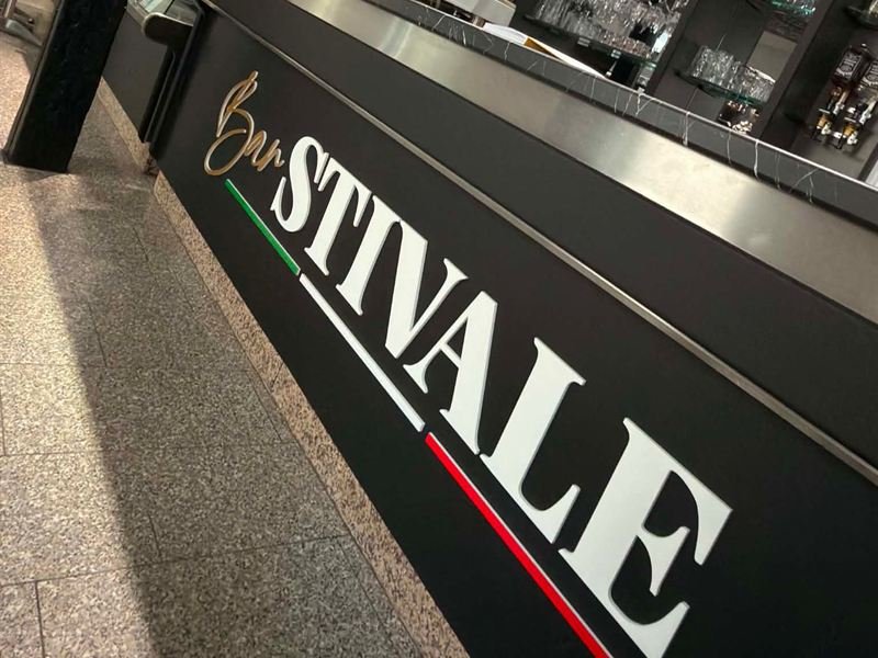 Die Thekenfront der Bar Stivale mit dem großen weißen Schriftzug Stivale auf schwarzem Grund, darüber die Marmorplatte und das Glasregal.