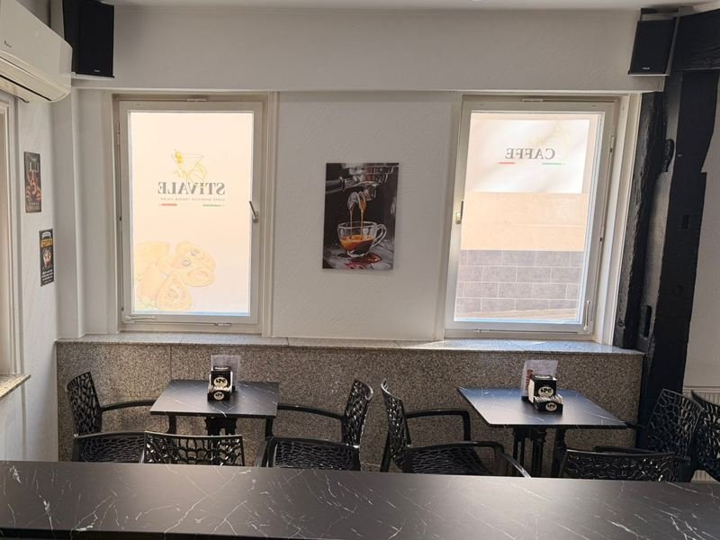 Blick von der Theke in den Gastraum der Bar Stivale: zwei Fenster mit Stivale- und Caffè-Folie, dazwischen ein Bild vom Espresso-Auslauf, davor Bistrotische aus schwarzem Marmor.