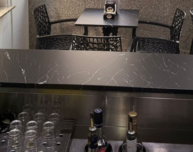 Blick von der Arbeitsseite der Bar über die schwarze Marmortheke: unten Gläser und Flaschen im Edelstahlbecken, dahinter ein Bistrotisch mit vier Stühlen.