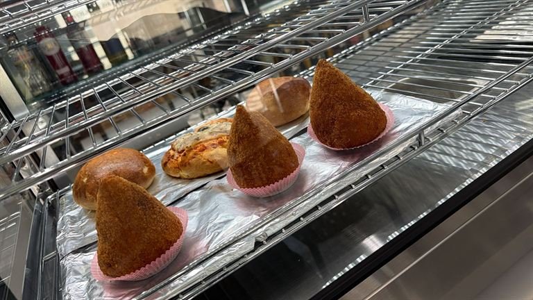 Blick in die warme Vitrine der Bar Stivale: vier Arancini in rosa Papierförmchen, dazwischen zwei Bombe und eine belegte Pizzette auf Alufolie.