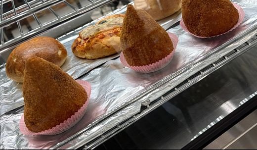 Nahaufnahme aus der warmen Vitrine: drei Arancini in rosa Papierförmchen, dahinter eine belegte Pizzette und zwei Bombe.